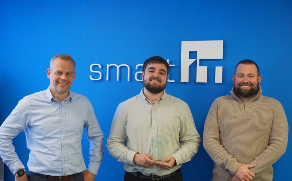 Three smiling men standing in front of Smart IT logo on blue wall.